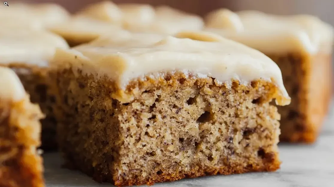 Close-up of a moist banana cake square topped with creamy vanilla frosting on a marble surface.