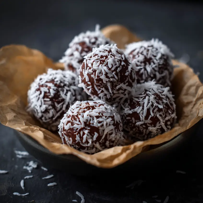 Bowl of rich chocolate coconut truffles generously coated in white shredded coconut, resting on a dark rustic surface.