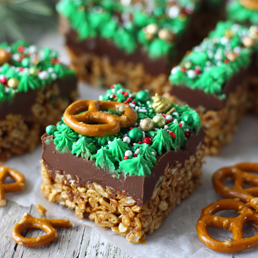 Close-up of festive three-layer No bake Christmas dessert bars with green frosting, sprinkles, and a mini pretzel on a wooden surface.
